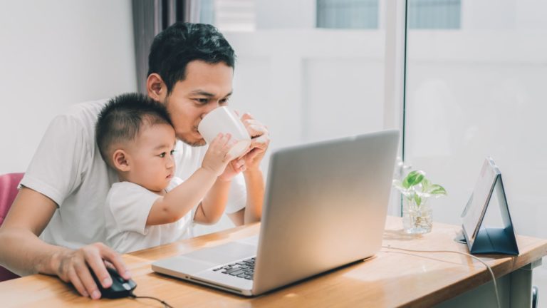 asian man holding his baby and a coffee cup looking at a laptop