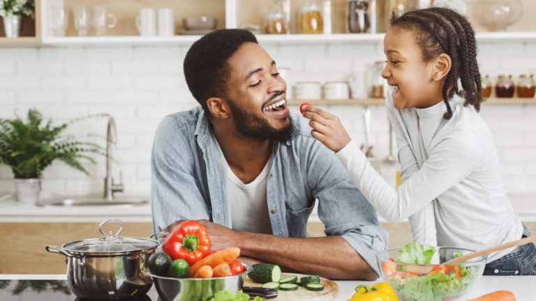 Cute african girl giving her dad cherry tomato while cooking