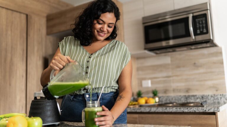 Healthy young woman making green smoothie in kitchen
